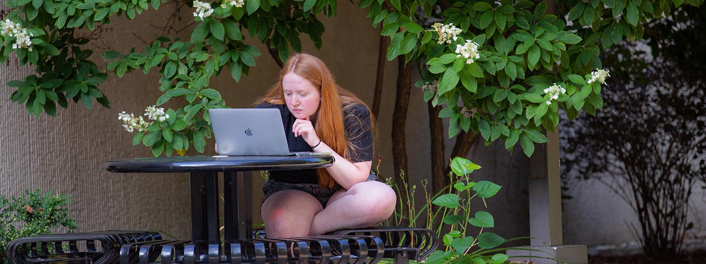 A student sitting at an outside table works on a laptop.