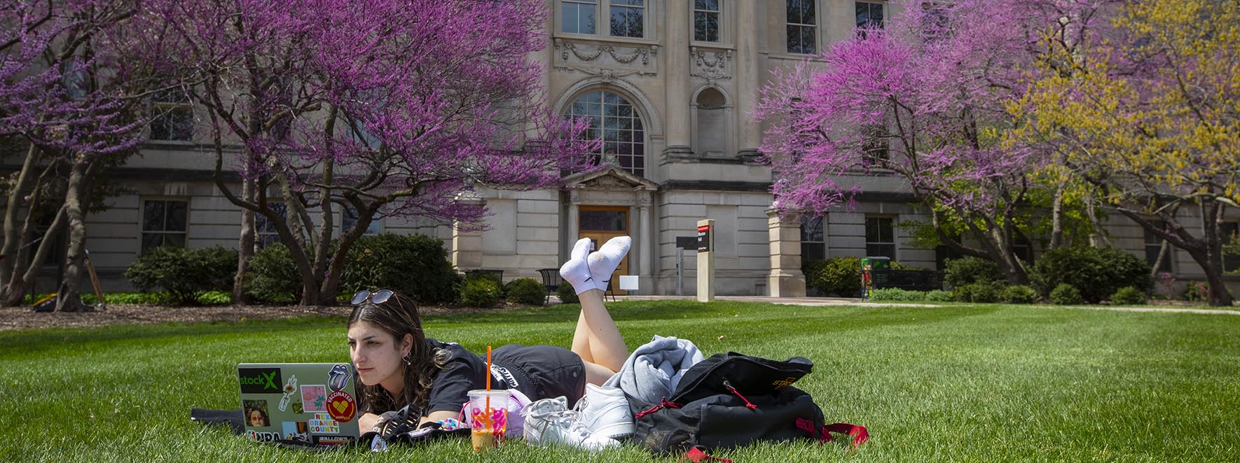A student works on a computer while laying on the grass outside Marston Hall