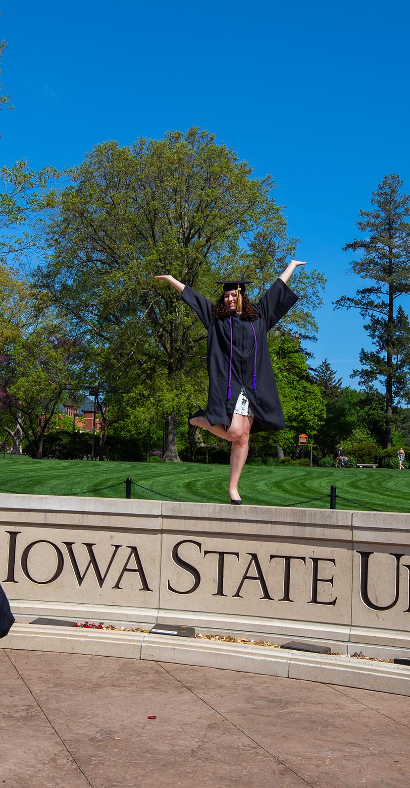 A graduate poses on the Iowa State University wall in front of the campanile