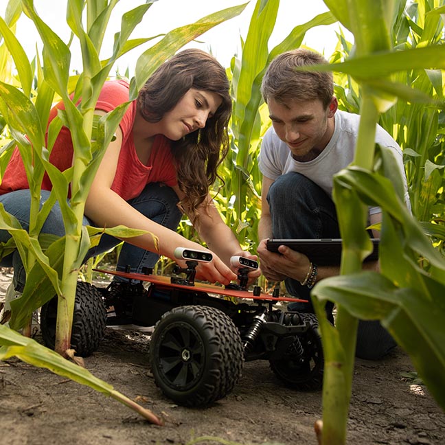 Two students crouch with a remote control exploratory vehicle in a corn field