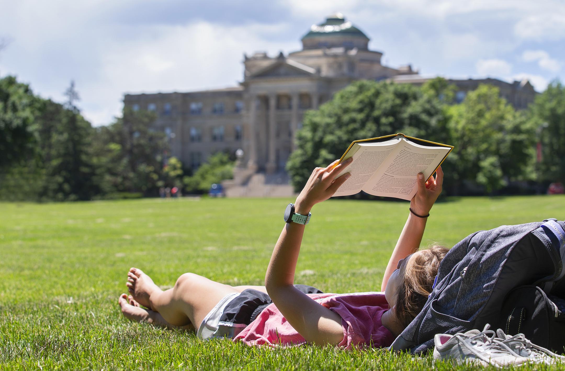 Student laying on sunny central campus reading a book with Beardshear Hall in the background