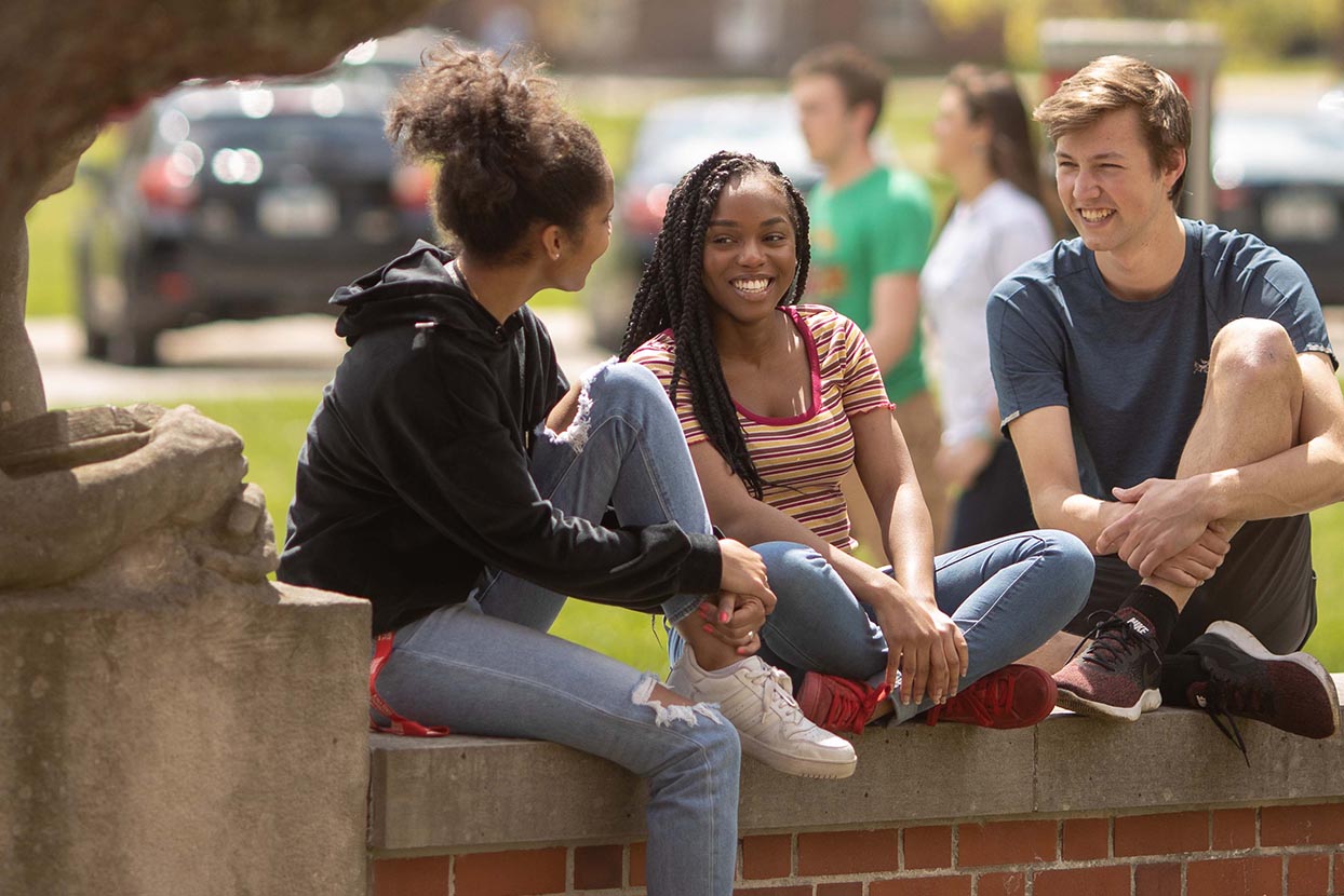 Three students talk while sitting on a brick wall