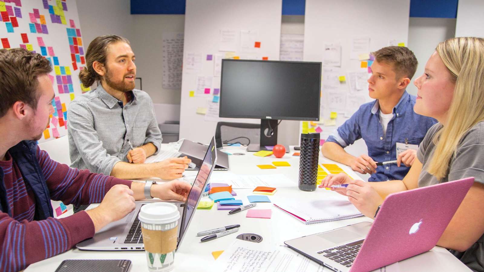 Four students meet at a table in a design lab surrounded with computers and project notes